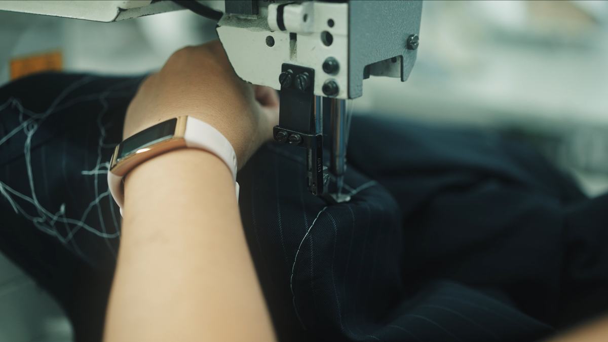 Sous l’aiguille d’une machine à coudre industrielle, une main guide un tissu marine à fines rayures