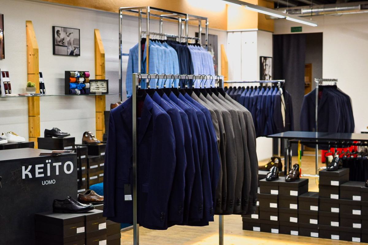 Des portants de vestes bleu marine et grises, des piles de boîtes à chaussures, le nom KEÏTO UOMO sur un meuble noir