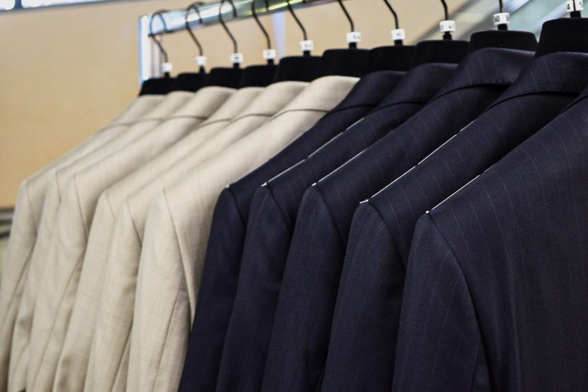 Une rangée de vestons sur cintres, épaules au premier plan, des beiges aux rayures bleu nuit