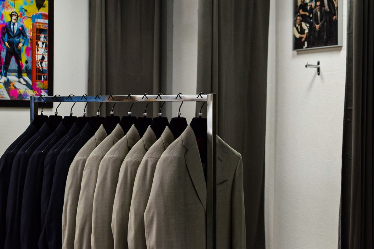 Un portant de vestons dans la boutique, des bleu nuit puis des beiges alignés sur leurs cintres, devant un rideau gris