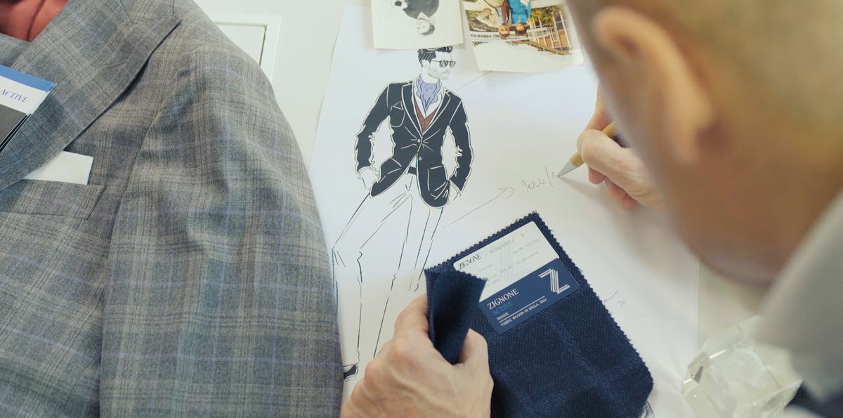 Deux mains au-dessus d’une grande feuille : l’une annote au crayon à côté du dessin imprimé d’un homme en veste sombre, l’autre présente un échantillon de laine bleue
