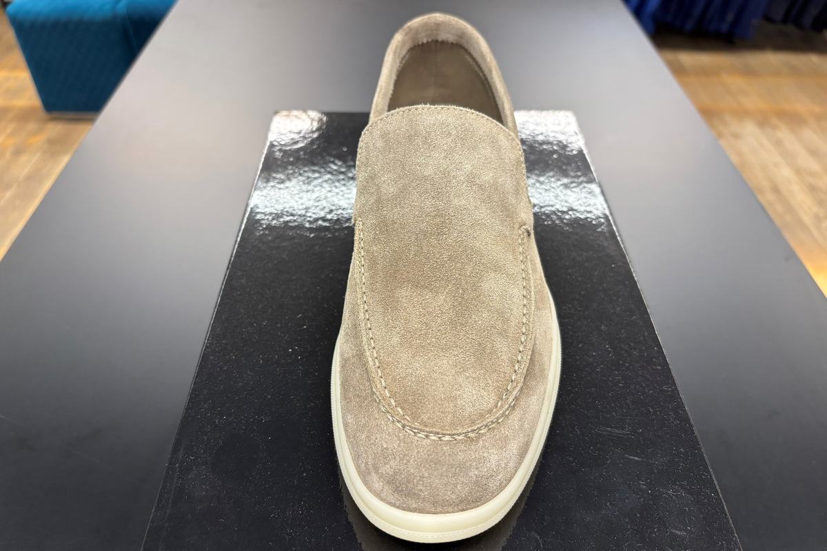 Un mocassin de daim taupe, semelle de caoutchouc crème, posé de face sur son coffret noir