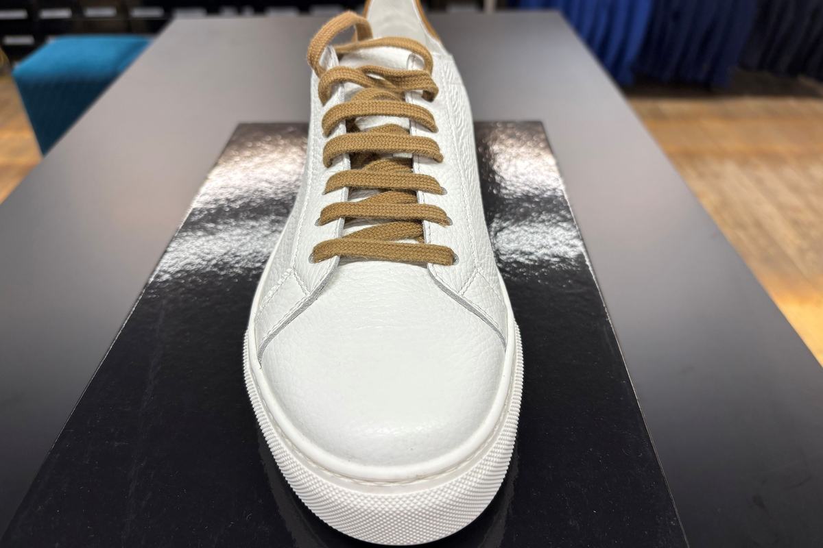 Une sneaker de cuir blanc grainé à lacets camel, semelle de caoutchouc blanc, posée de face sur son coffret noir