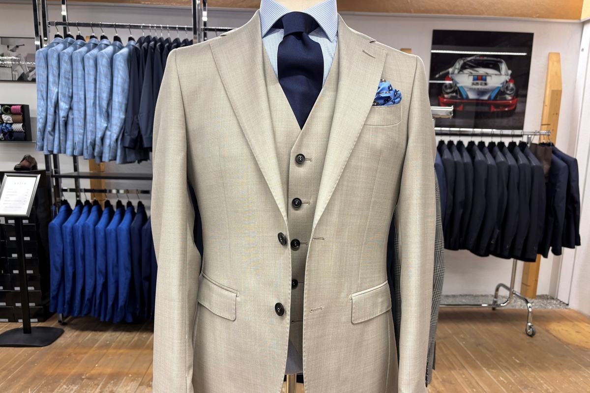 Un costume trois-pièces de laine sable sur mannequin, gilet boutonné, chemise bleu clair, cravate marine et pochette de soie bleue